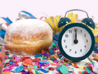 Krapfen mit einer Uhr, die auf 12 steht, Luftschlangen im Hintergrund. Faschingsstimmung.