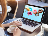 Frau sitzt mit Tasse Cappucchino vor Laptop in einem Kaffee und auf dem Bildschirm sind bunte Donuts auf hellblauem Hintergrund zu sehen