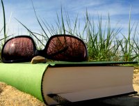 Buch und Sonnenbrille im Sand, im Hintergrund Gras