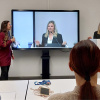 Studierende üben einen Vortrag im Übungspresentationsraum. Eine große Leinwand ist sichtbar, auf der sich eine Sprecherin spiegelt und Publikum, das zuhört, ist im Vordergrund zu sehen
