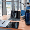 Lernplatz mit Laptop, Schreibblock, Stift, Tablet und Trinkfalsche in der Bibliothek mit einem Studierenden und einem Bücherregal im Hintergrund