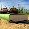 Buch und Sonnenbrille im Sand, im Hintergrund Gras