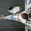 Student sits on floor in library leafs through notebook, laptop and bookshelf in background