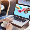 Frau sitzt mit Tasse Cappucchino vor Laptop in einem Kaffee und auf dem Bildschirm sind bunte Donuts auf hellblauem Hintergrund zu sehen